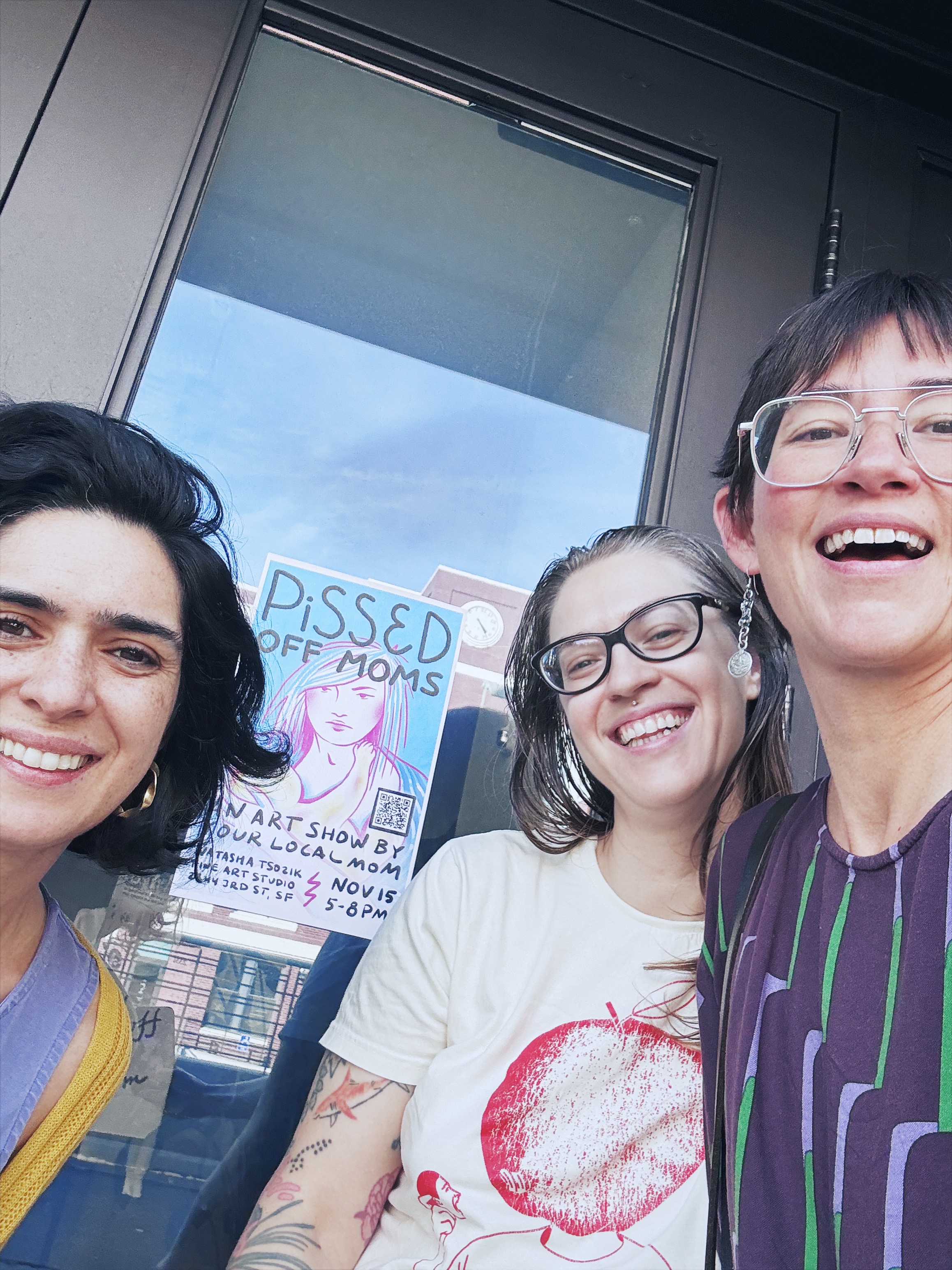 Three friends smiling outside the studio window, the POM 2.0 poster behind them.
